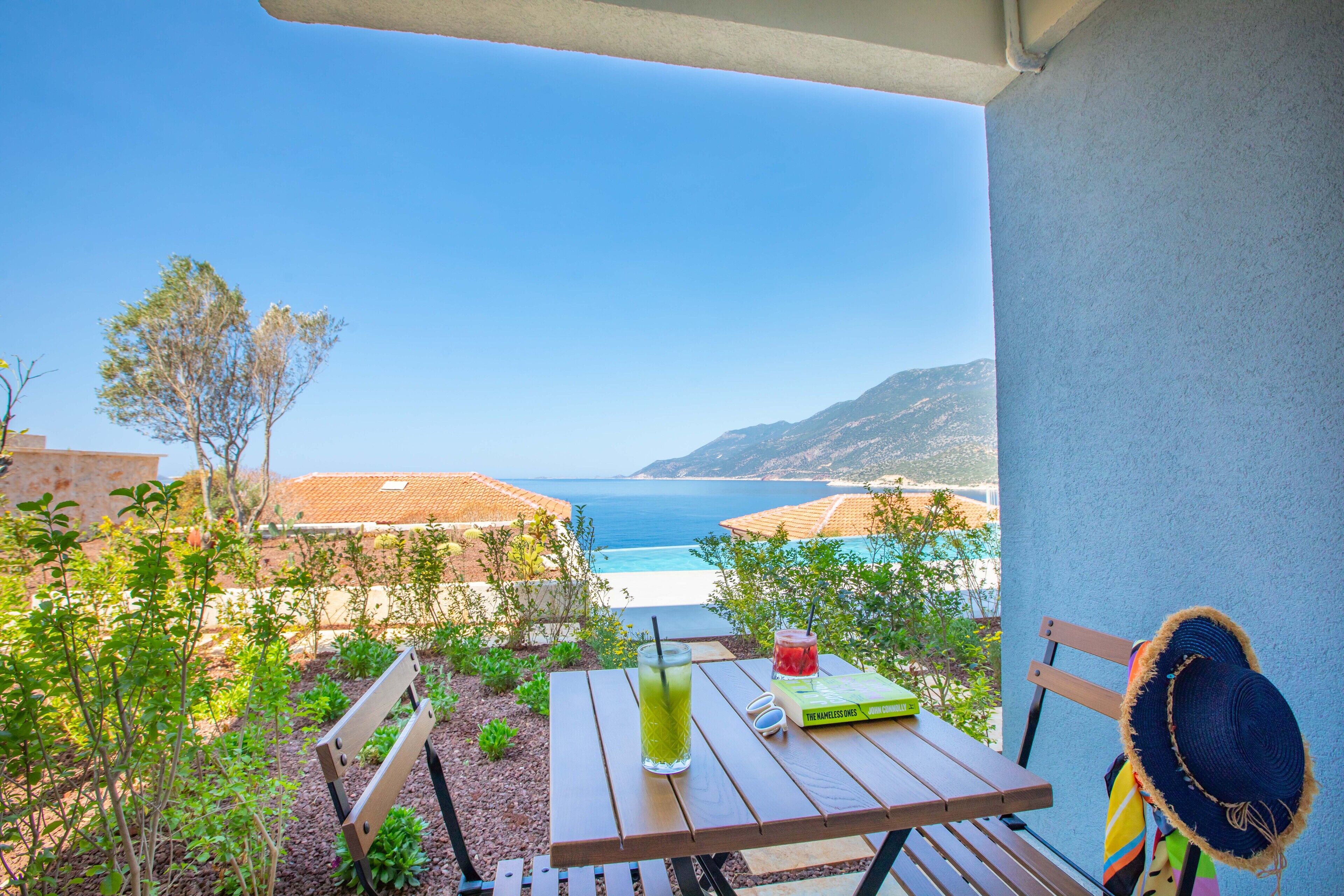 A wooden table with a green drink, a book, and a hat on a balcony overlooking a beach and mountains.