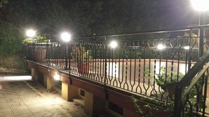 Une vue nocturne d’une clôture métallique illuminée, d’un chemin dallé et d’un bâtiment avec un toit rouge.