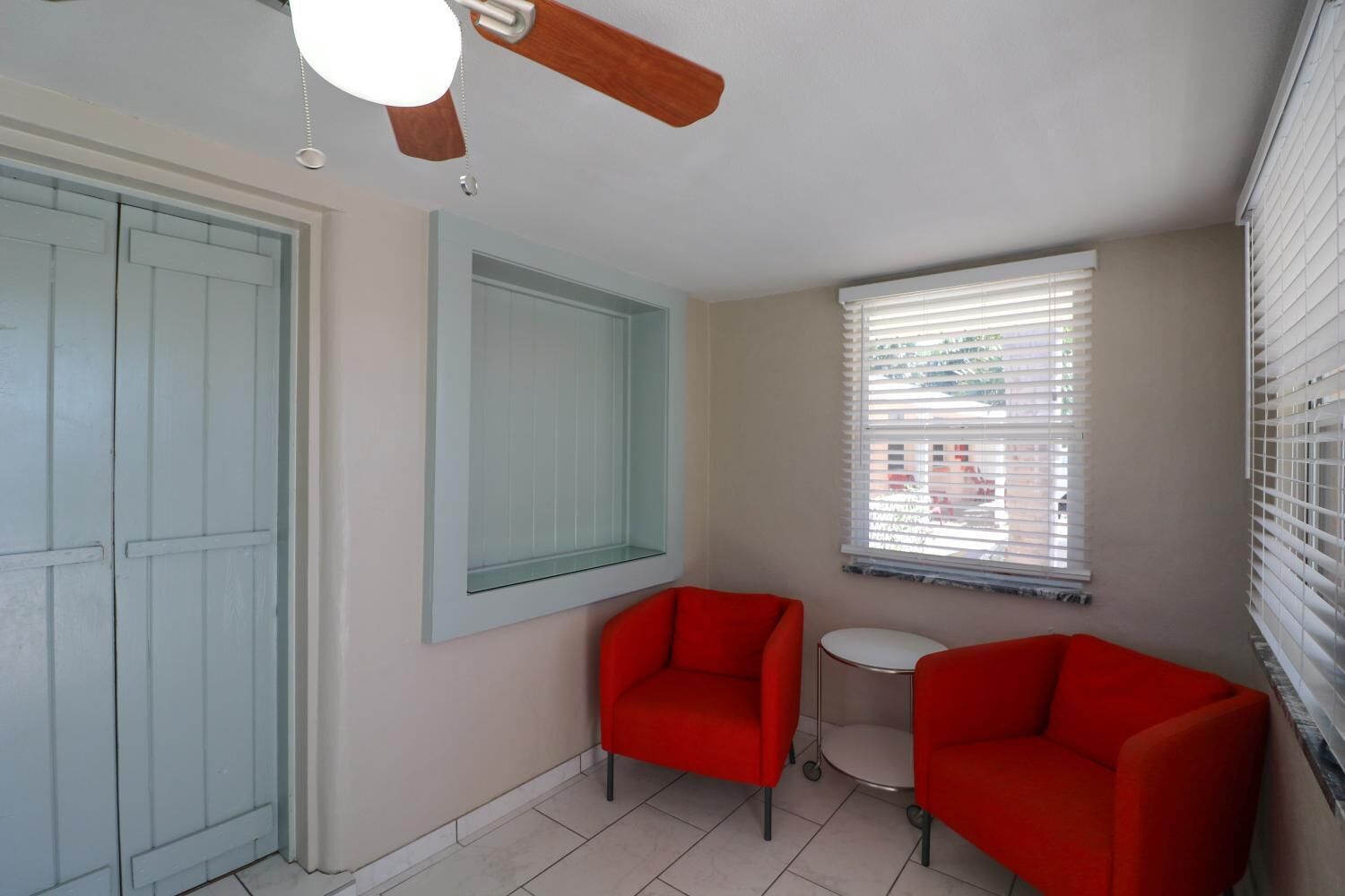 Una habitación con ventilador de techo, ventana con persianas, dos sillas rojas y una pequeña mesa redonda.