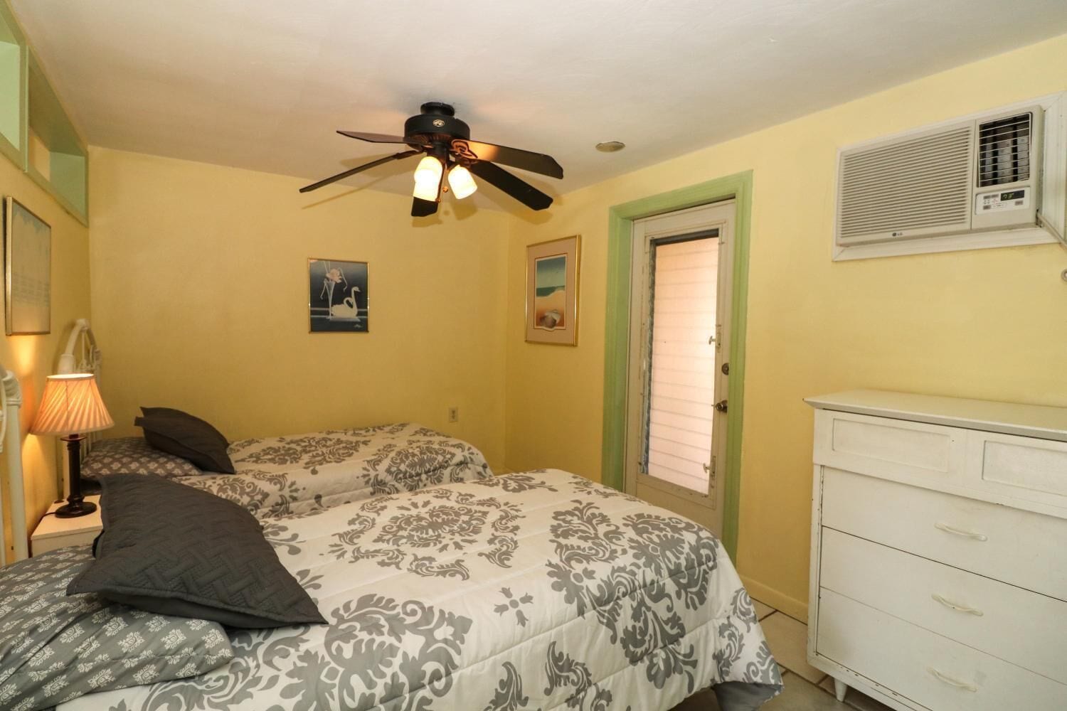 A bedroom with two beds, a ceiling fan, a bedside lamp, a dresser, and an air conditioning unit.