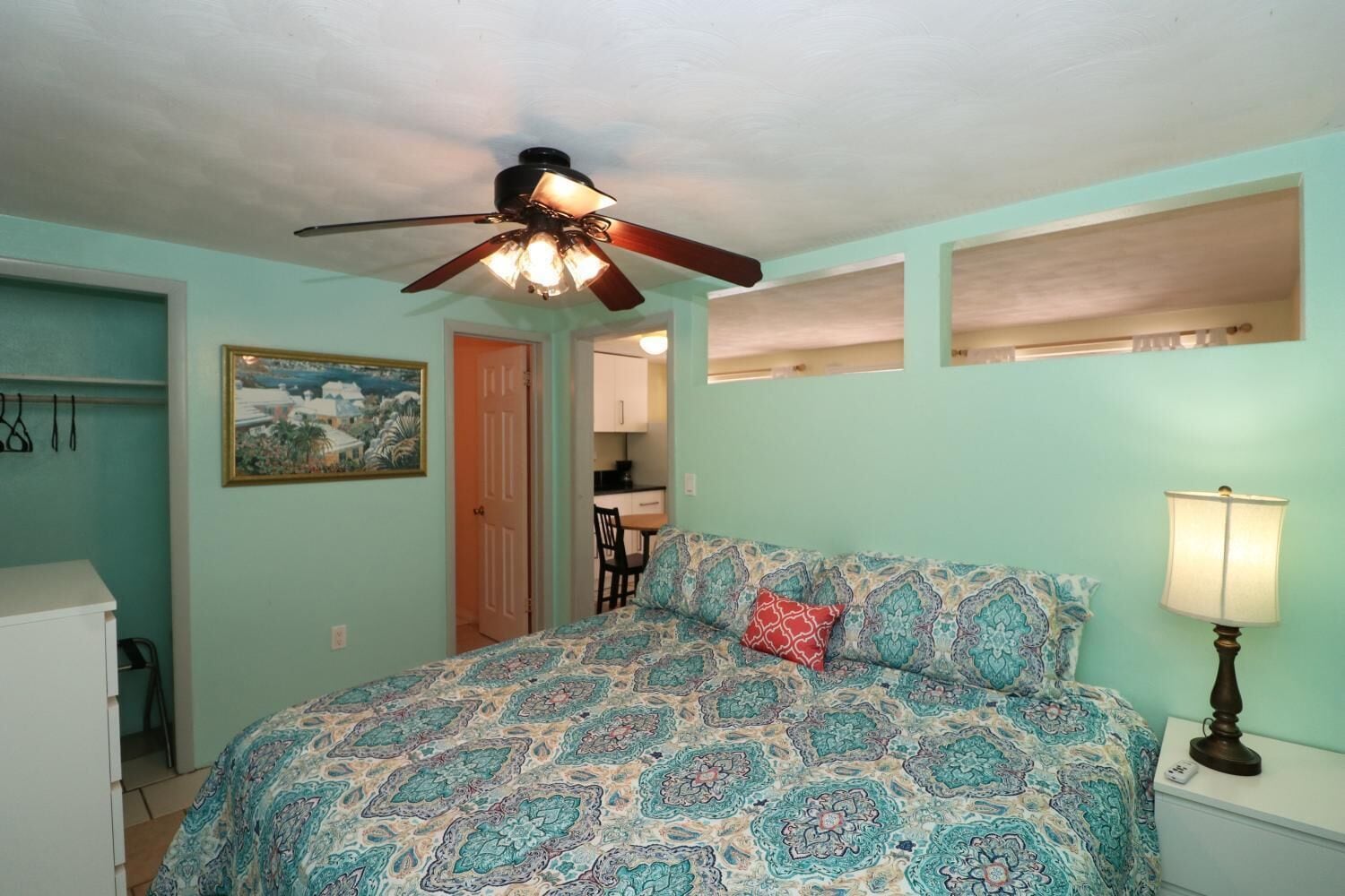 A bedroom with a bed, a ceiling fan, a nightstand with a lamp, a painting on the wall, and a door leading to a bathroom.
