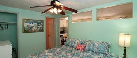 A bedroom with a bed, a ceiling fan, a nightstand with a lamp, a painting on the wall, and a door leading to a bathroom.