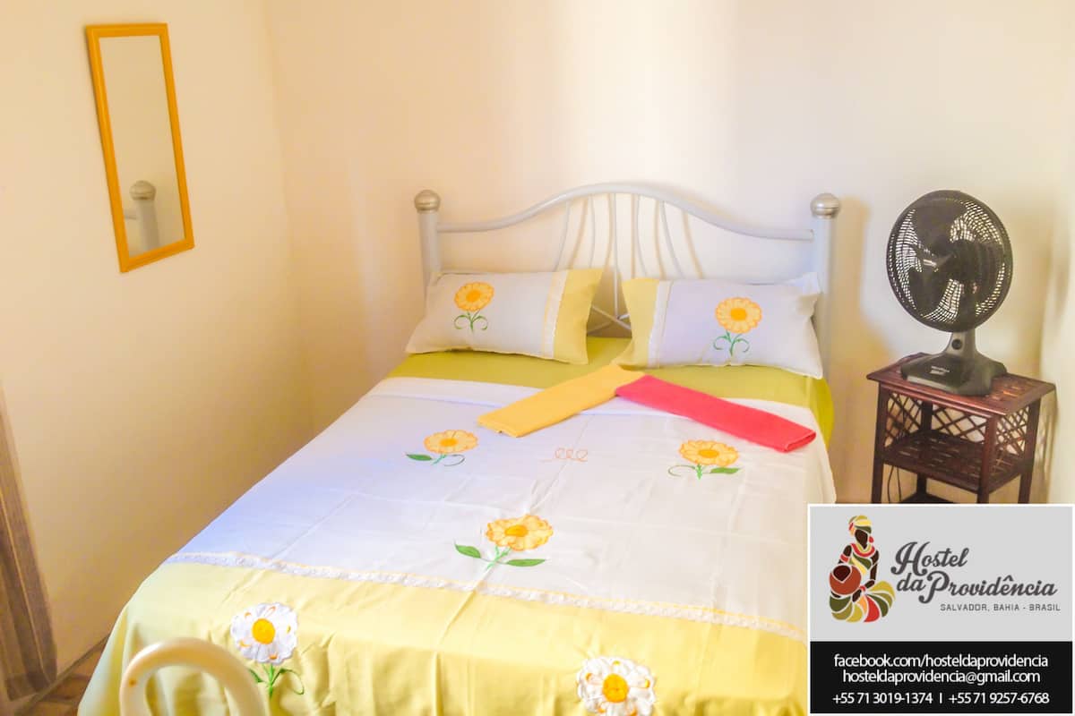 A bed with a white headboard, yellow and white floral bedding, and a bedside fan.