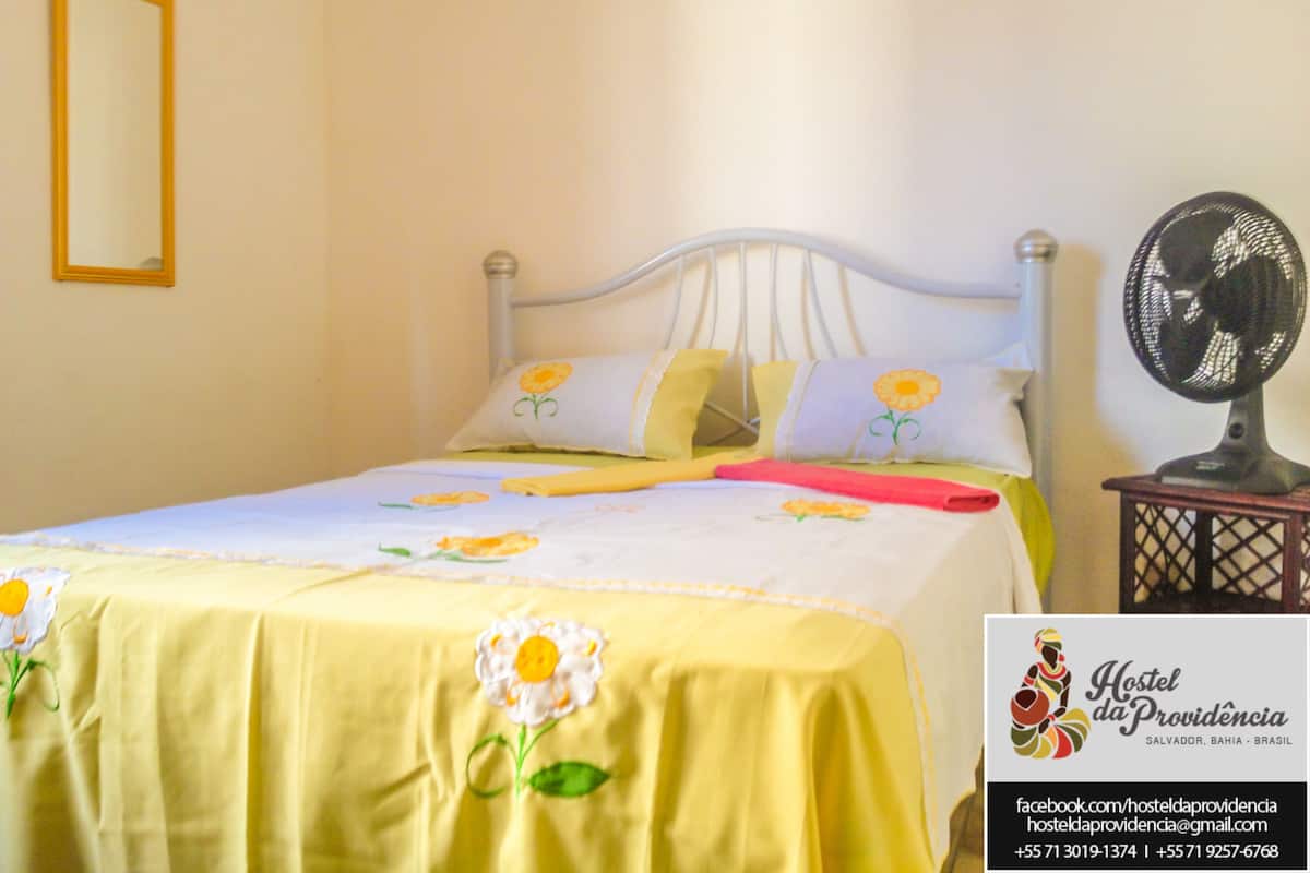 A bed with a yellow bedspread and white pillows with yellow flowers, a bedside fan, and a mirror on the wall.