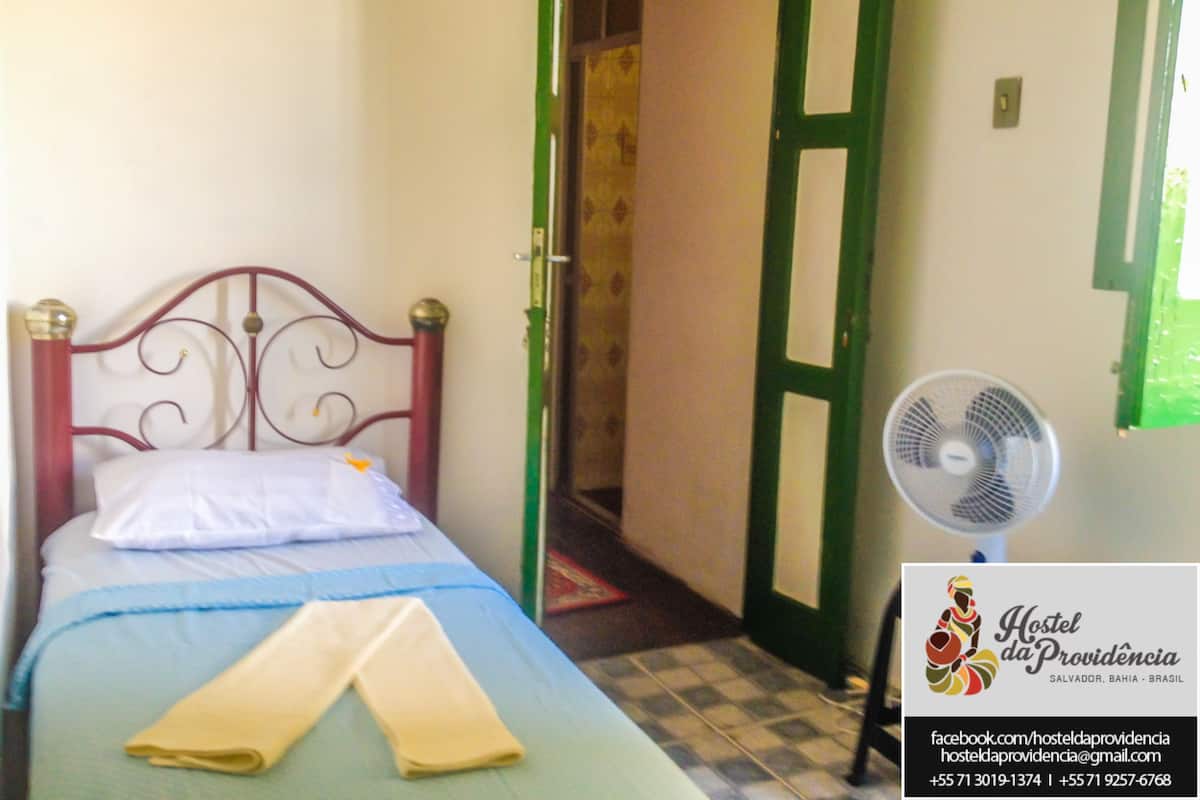 A bed with a metal headboard, a white pillow, and a blue bedspread. There is a folded towel on the bed. A standing fan is next to the bed.