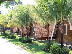A row of wooden cabins with thatched roofs, surrounded by lush greenery and palm trees. - Pousada Monte Carlo (Olímpia)