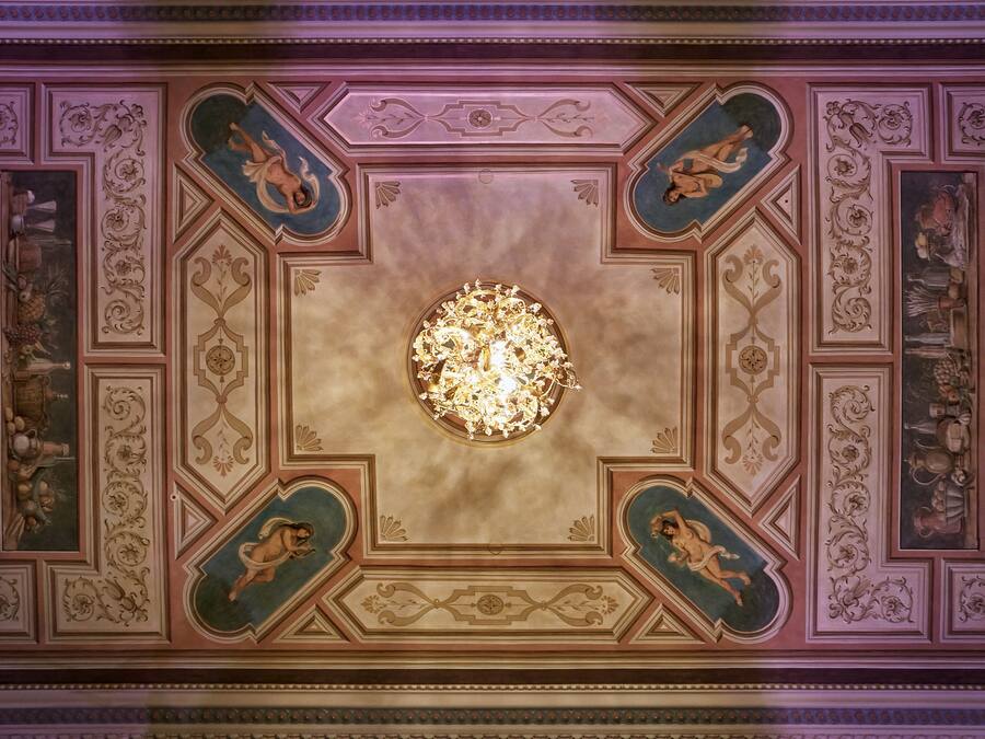 A ceiling with intricate floral and geometric patterns, a central chandelier, and framed artwork.