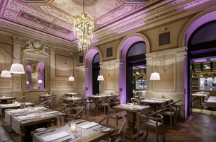 Un ristorante con soffitto decorato e lampadari, tavoli e sedie in legno, ed illuminazione con tocchi viola.