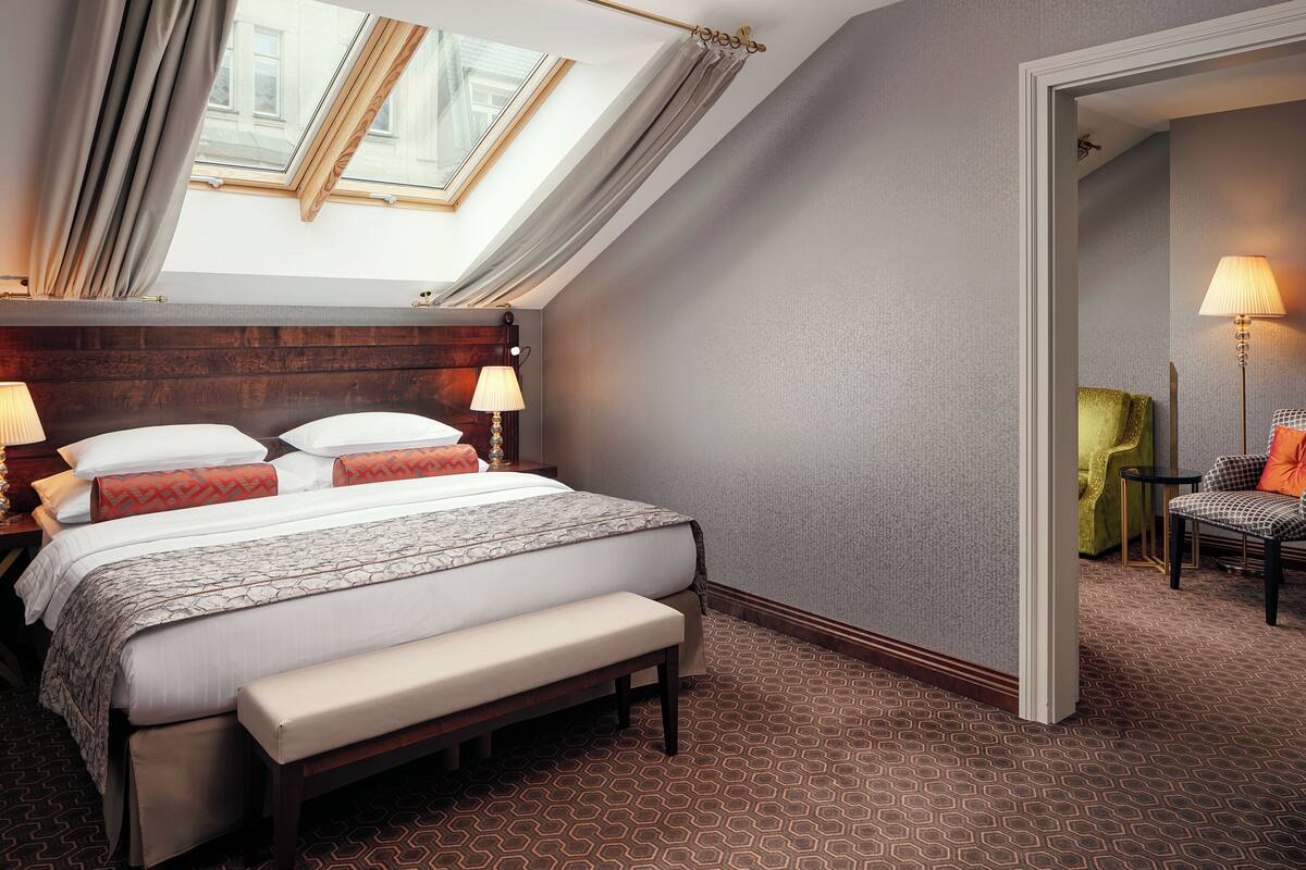 A hotel room with a large bed, a bench, a skylight, a lamp, and a chair.