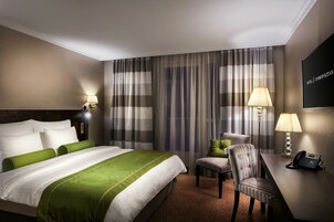 Quarto de hotel com uma cama grande, uma escrivaninha com telefone, uma cadeira e uma televisão na parede.