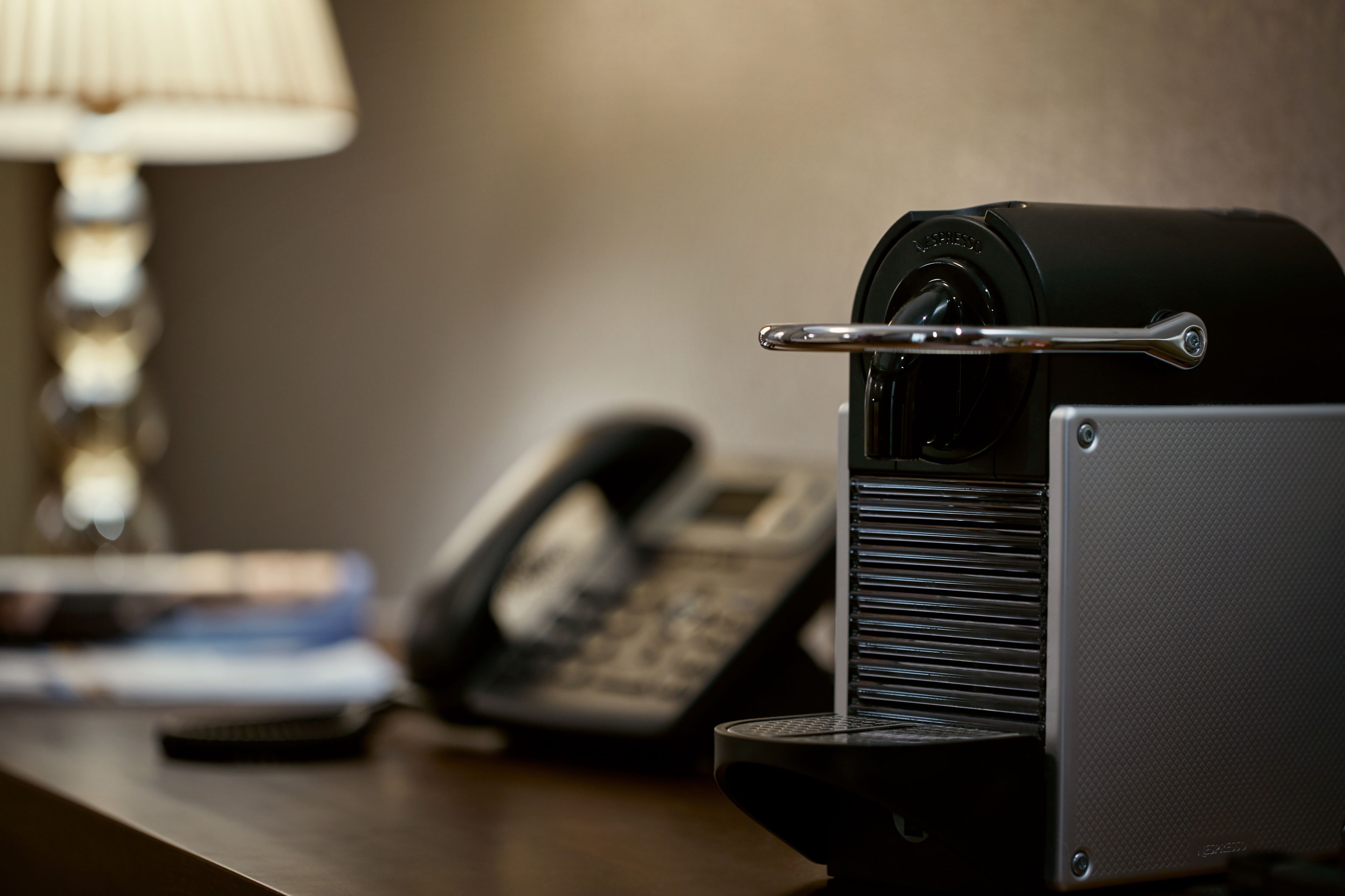 Una máquina de café sobre un escritorio, con un teléfono y una lámpara al fondo.