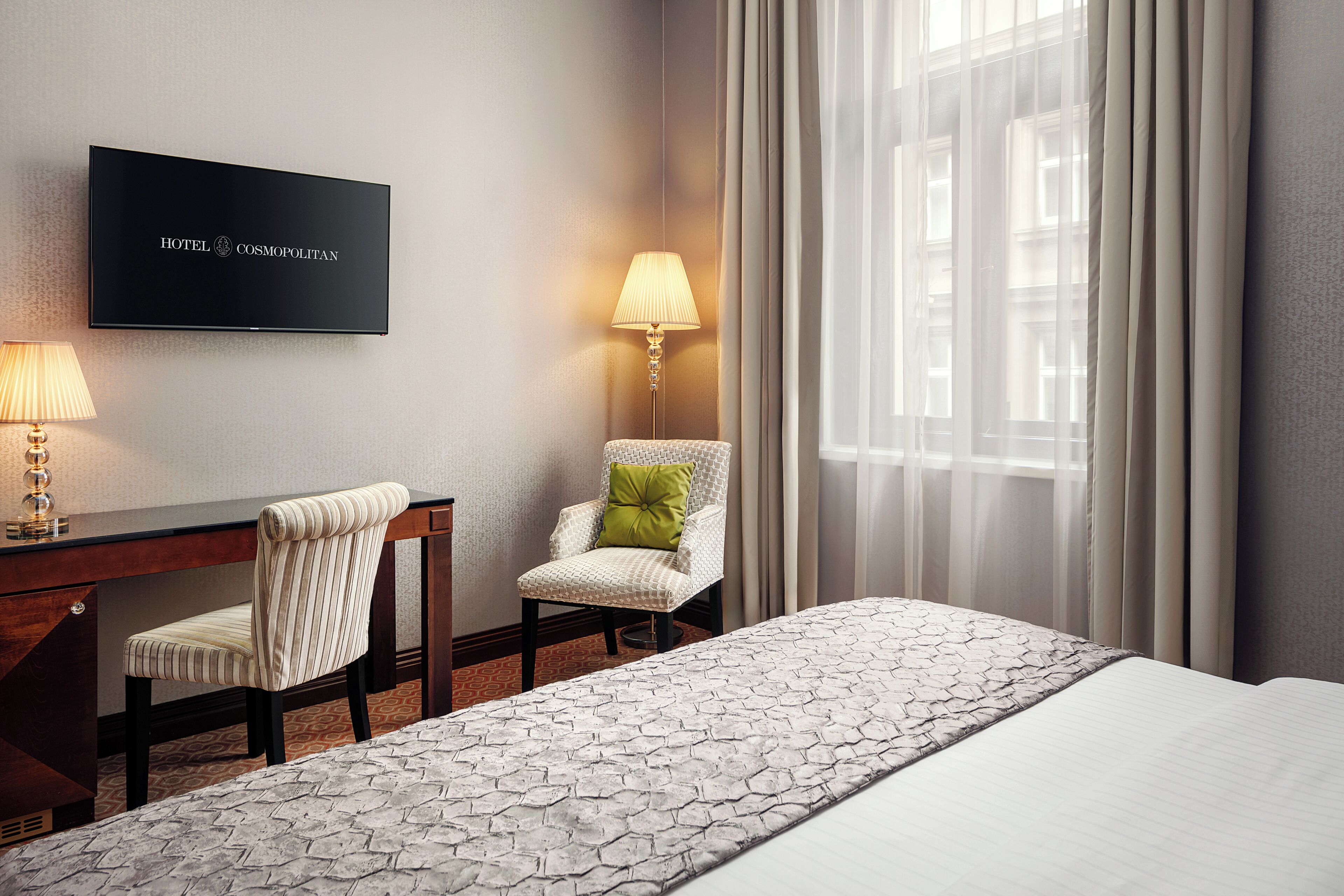 Una camera d'albergo con un letto, una scrivania, una sedia, una TV a muro e una finestra con tende.