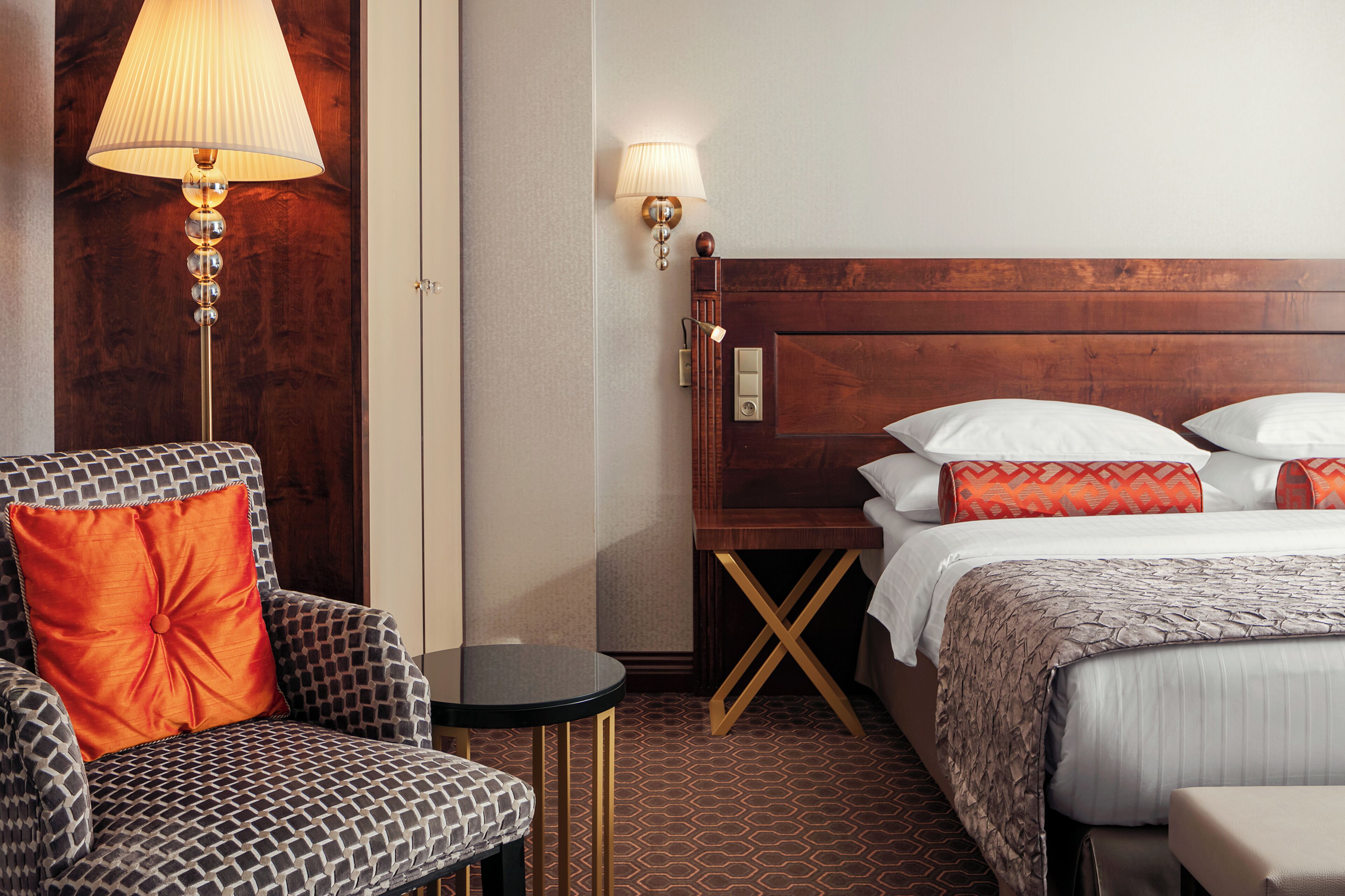 A hotel room with a wooden headboard, a patterned armchair, a small round table, and a bed with white and orange pillows.