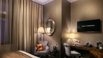 Una camera d'albergo con un letto, una scrivania con telefono e macchina del caffè, una sedia con cuscino, uno specchio e una televisione a parete.