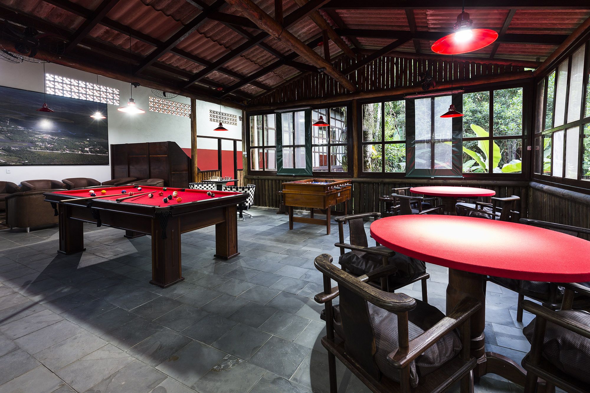 A pool table, foosball table, and billiard table in a room with large windows and red tables.