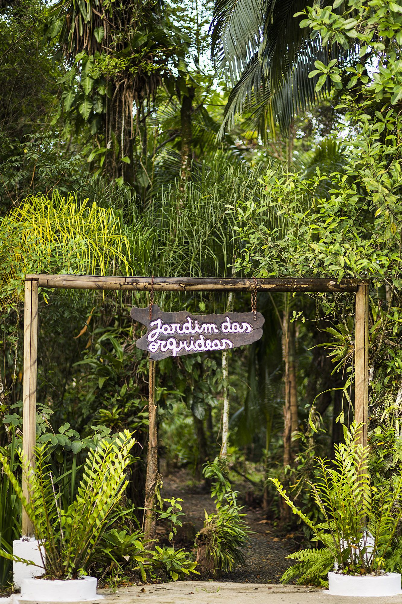 Uma pérgola de madeira com um letreiro escrito 'Jardim das ervidas', cercada por vegetação exuberante.