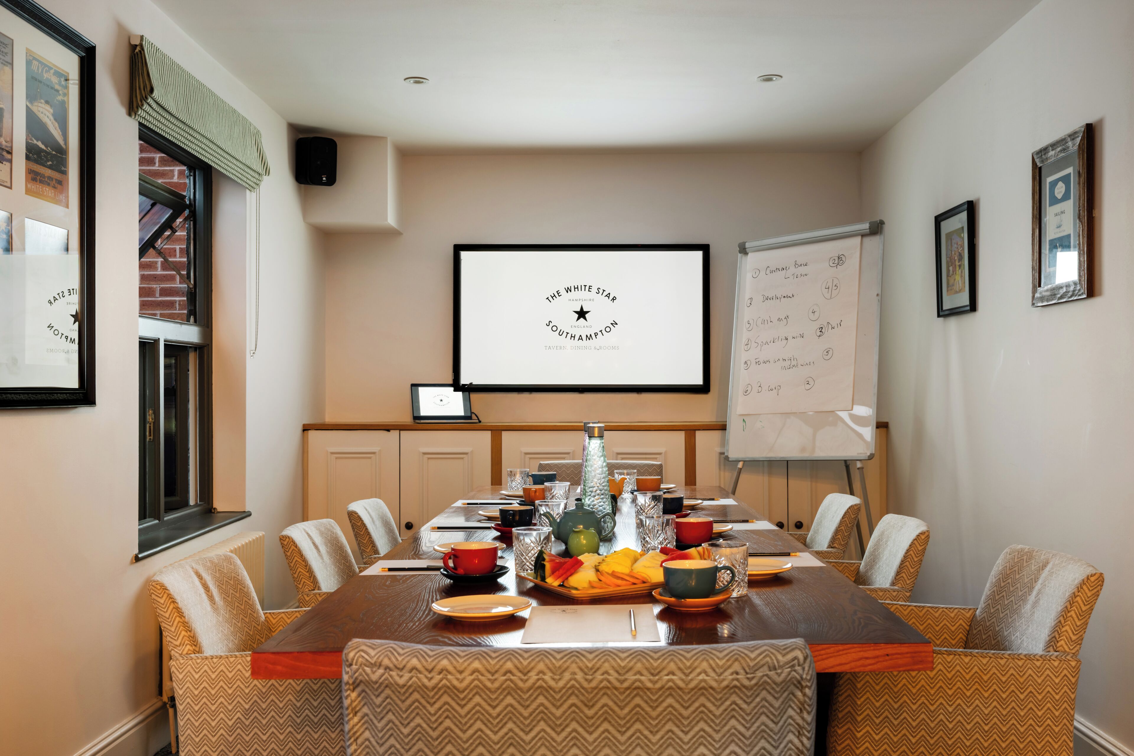 Uma sala de conferência com uma mesa comprida, cadeiras, uma tela de projetor, um quadro branco e um quadro na parede.
