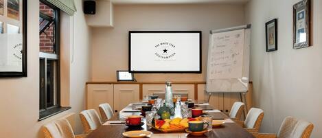 A conference room with a long table, chairs, a projector screen, a whiteboard, and a framed picture on the wall.