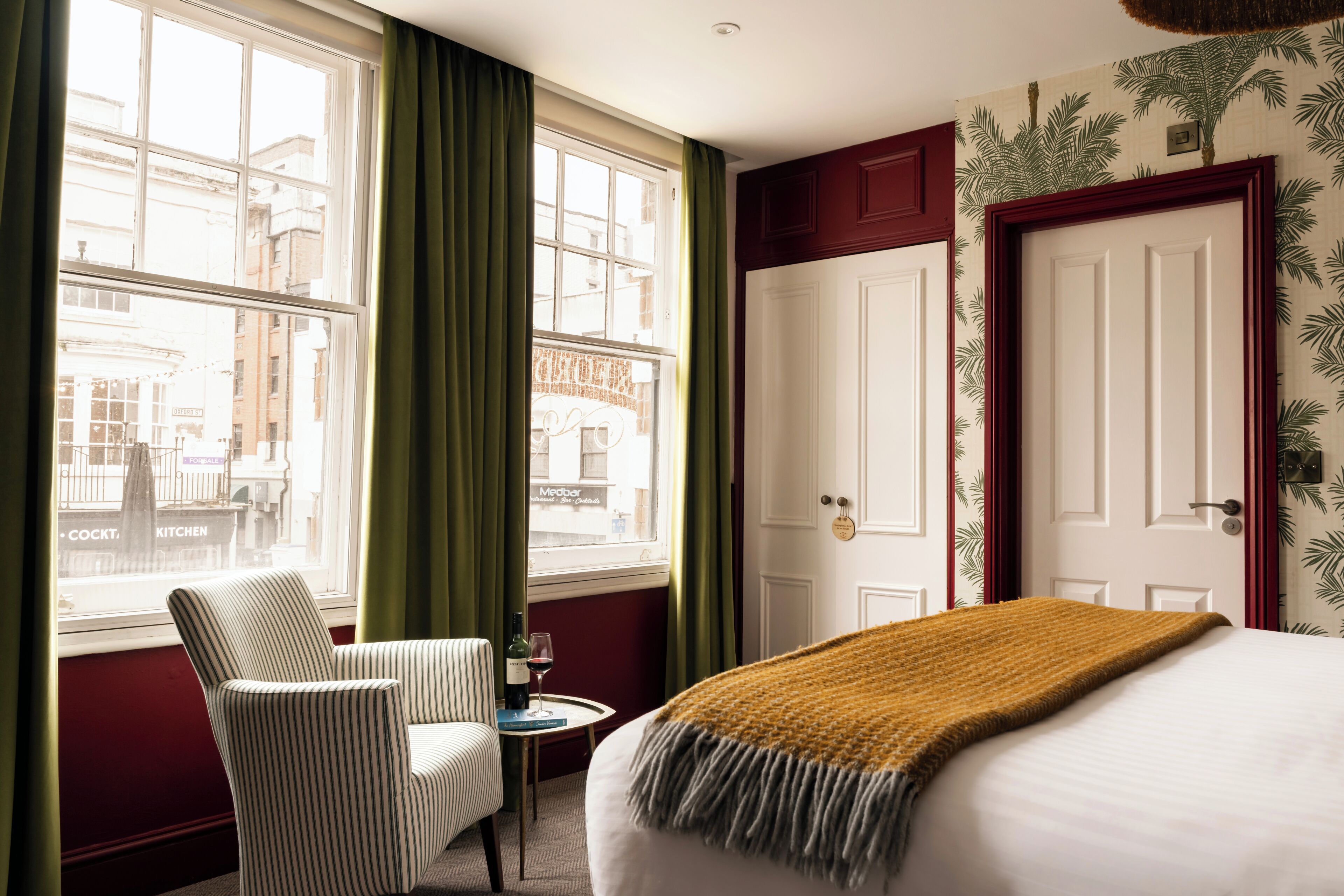 A hotel room with a bed, a chair, a small table with a wine glass, and a view of buildings through large windows.