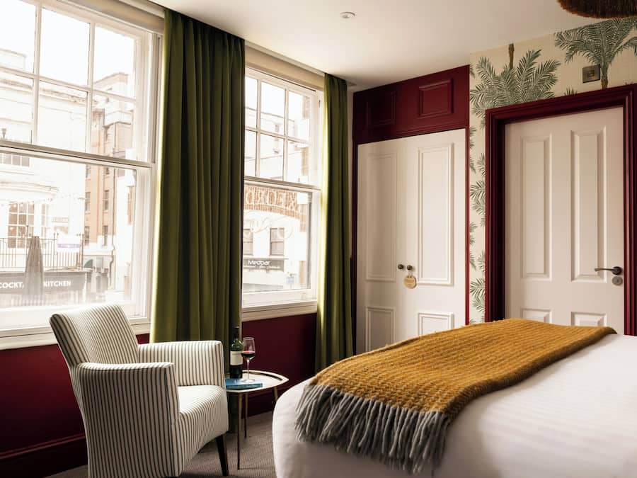 Habitación de hotel con cama, una silla, una mesita con una copa de vino y vista a edificios a través de grandes ventanales.
