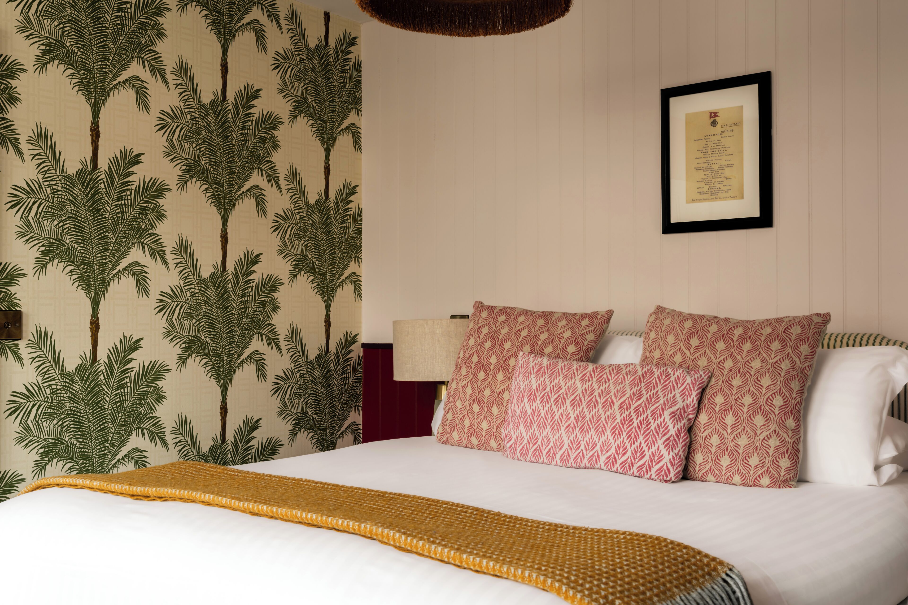 A neatly made bed with patterned pillows, a framed certificate on the wall, and a wall with a palm tree pattern.