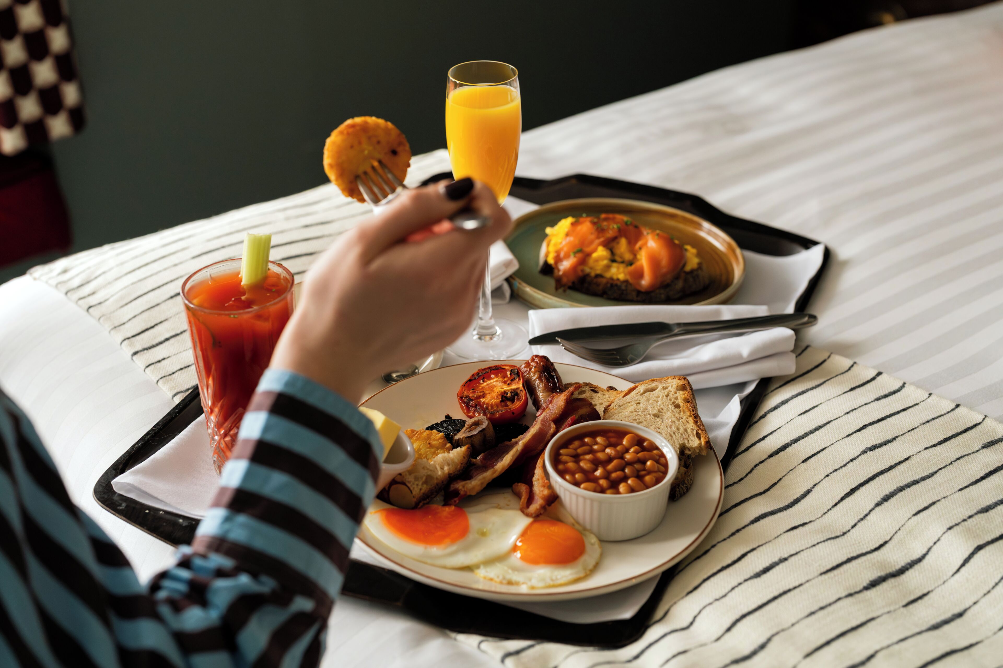 A breakfast tray with eggs, bacon, toast, beans, and a glass of orange juice on a bed.