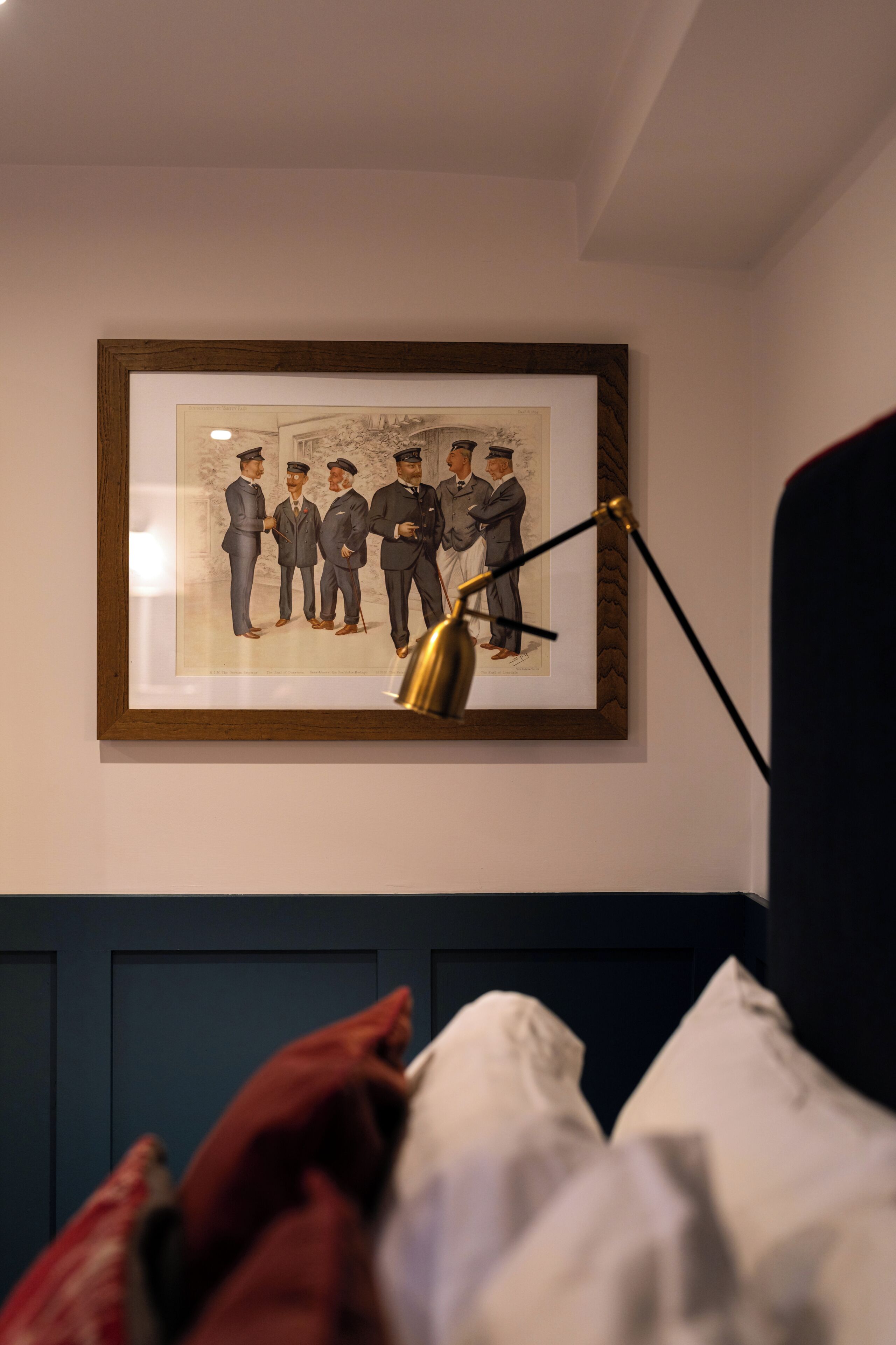 A framed picture of men in suits on a wall, a desk lamp, and a bed with a white headboard.