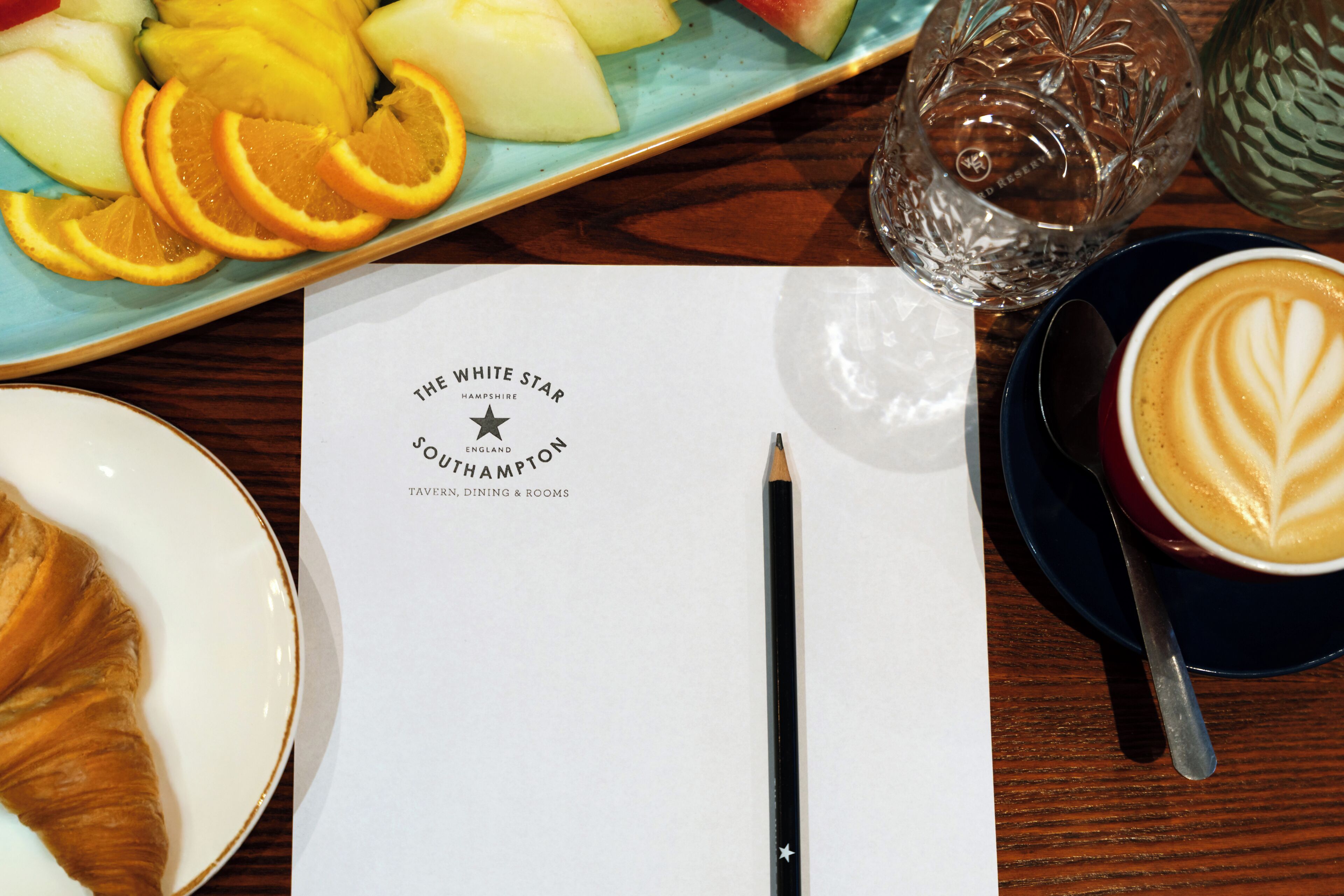 A table with a plate of fruit, a cup of coffee, a menu, and a pencil.