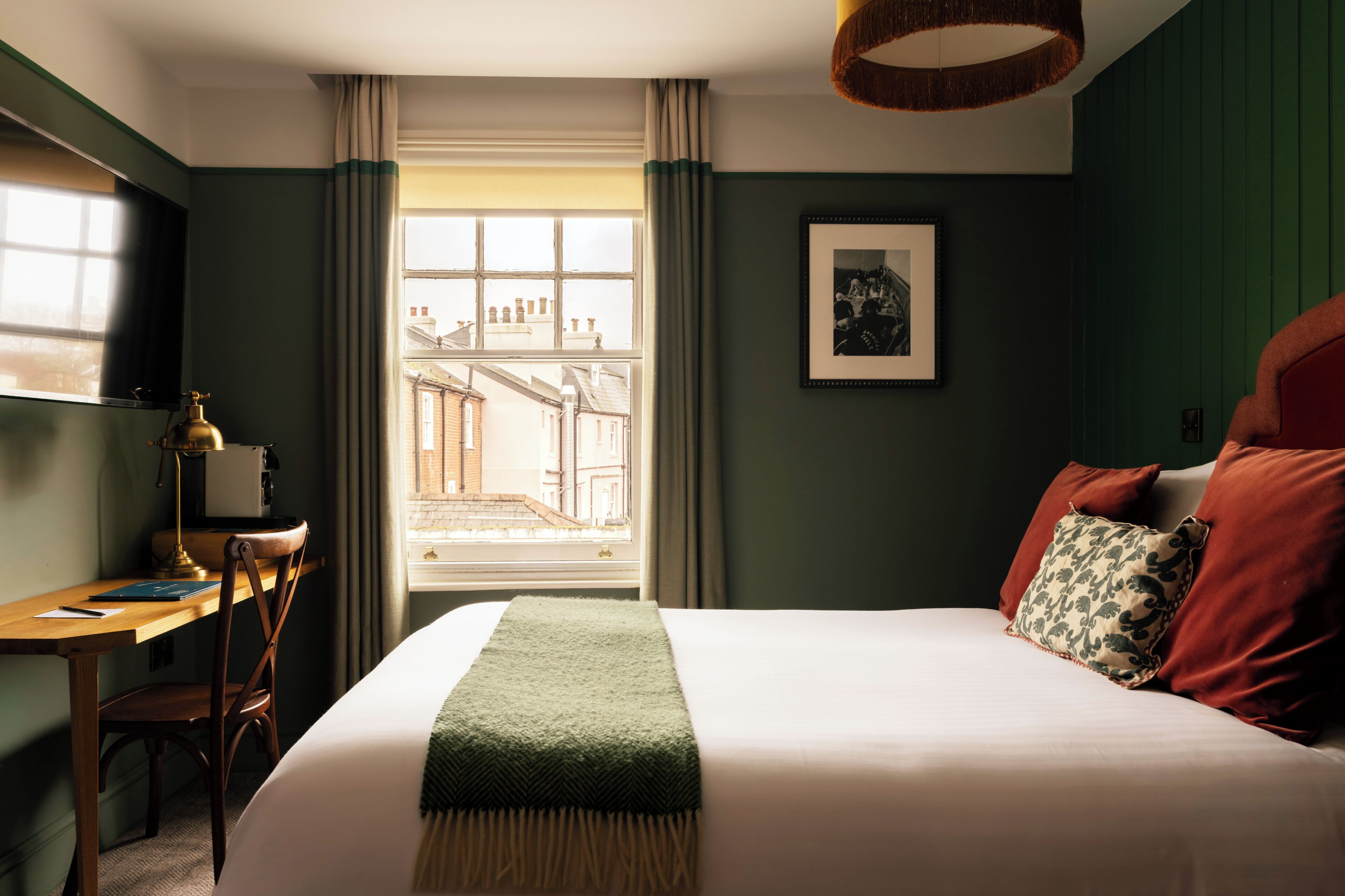 A bedroom with a bed, a desk, a chair, a window with curtains, and a framed picture on the wall.