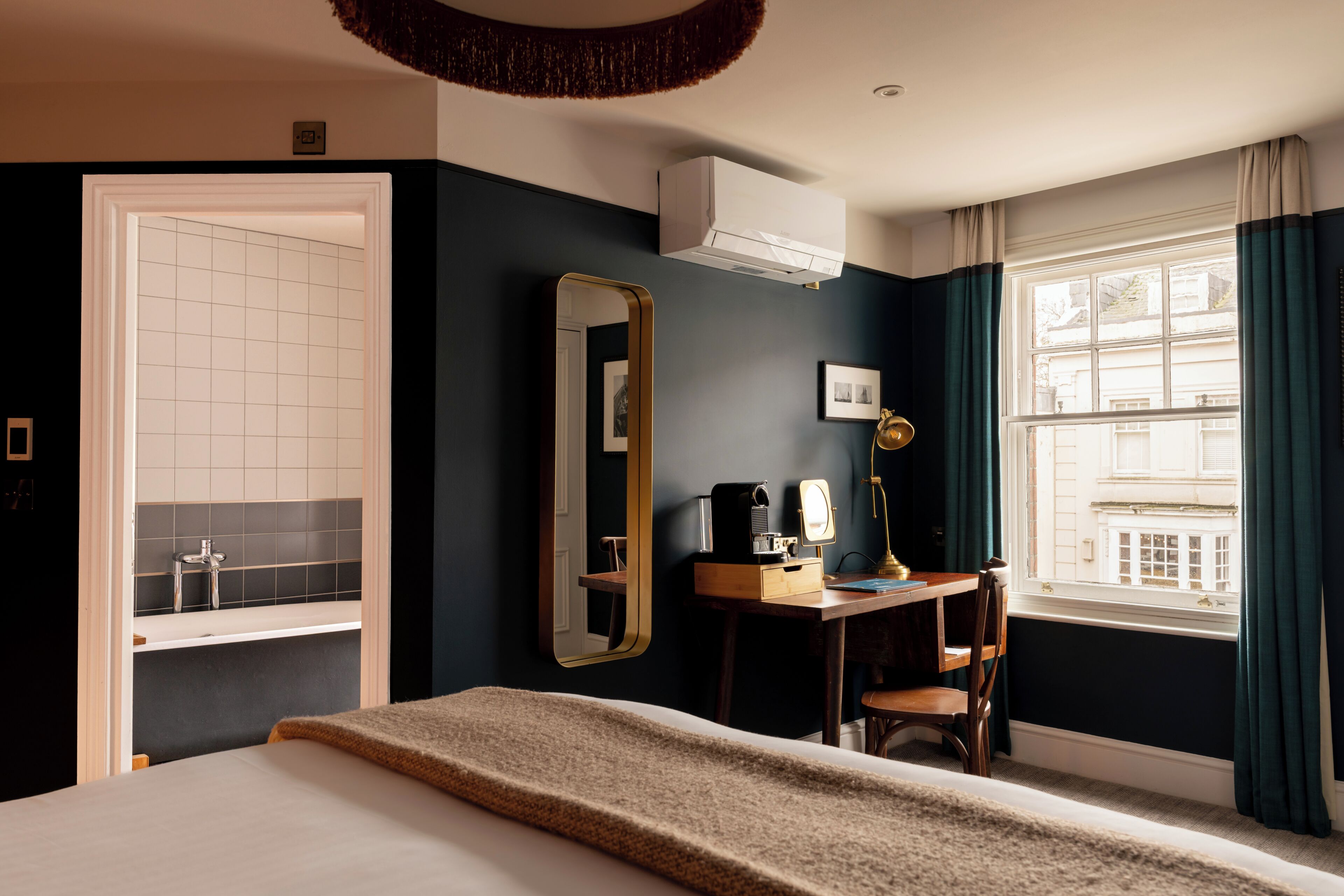 A hotel room with a bed, a desk with a lamp, a mirror, and a window with curtains.