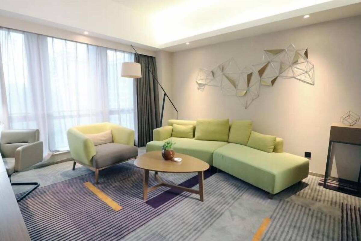 A modern hotel room with a green sofa, a small round table, a lamp, and abstract wall art.