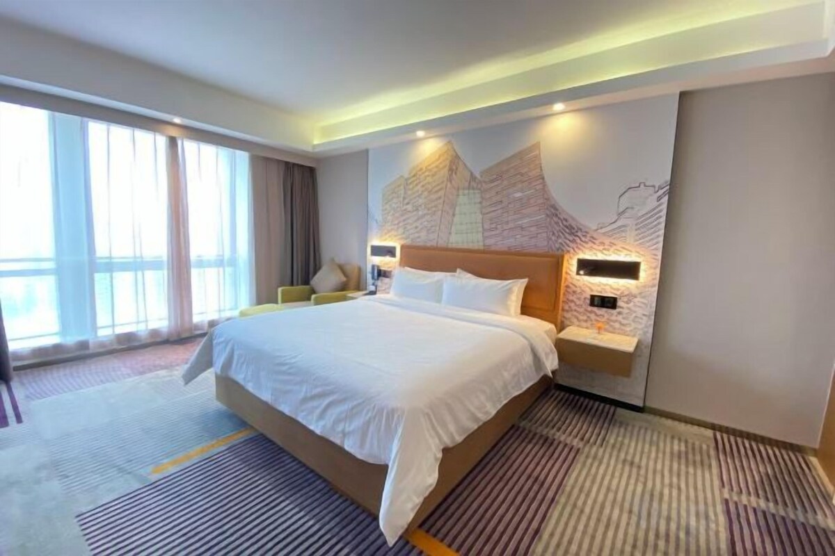 Habitación de hotel con una cama grande, una mesita de noche con lámpara, un televisor montado en la pared y un ventanal con cortinas.