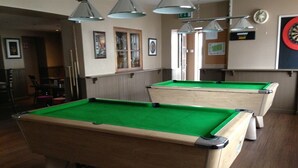 Una mesa de billar con superficie de fieltro verde, estructura de madera y un bolsillo trasero negro. Hay luces colgantes y un dardo en la pared.