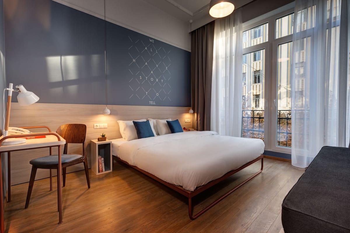 A modern hotel room with a large bed, a desk, a chair, and a view of the city through large windows.