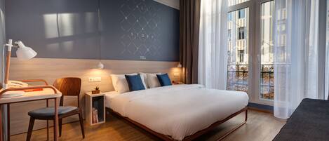 A modern hotel room with a large bed, a desk, a chair, and a view of the city through large windows.