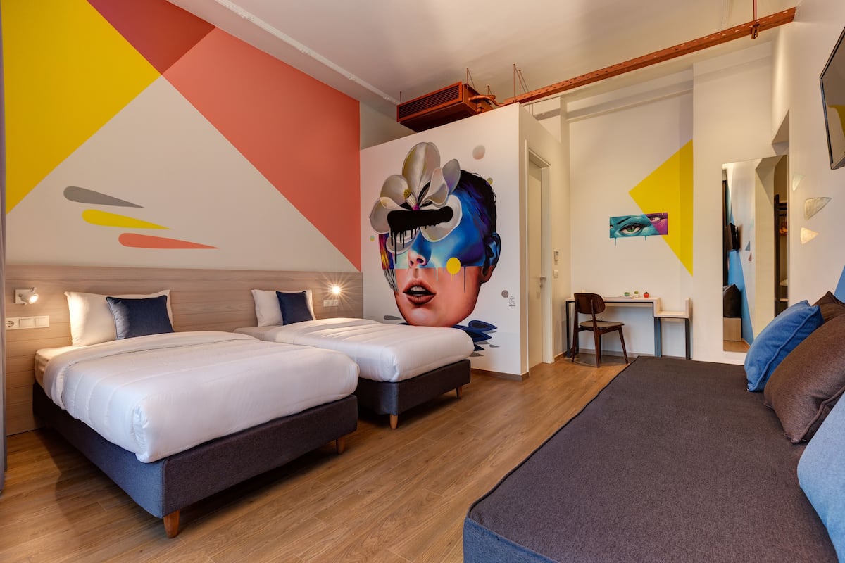 A modern hotel room with two beds, a desk, and a large mural on the wall.