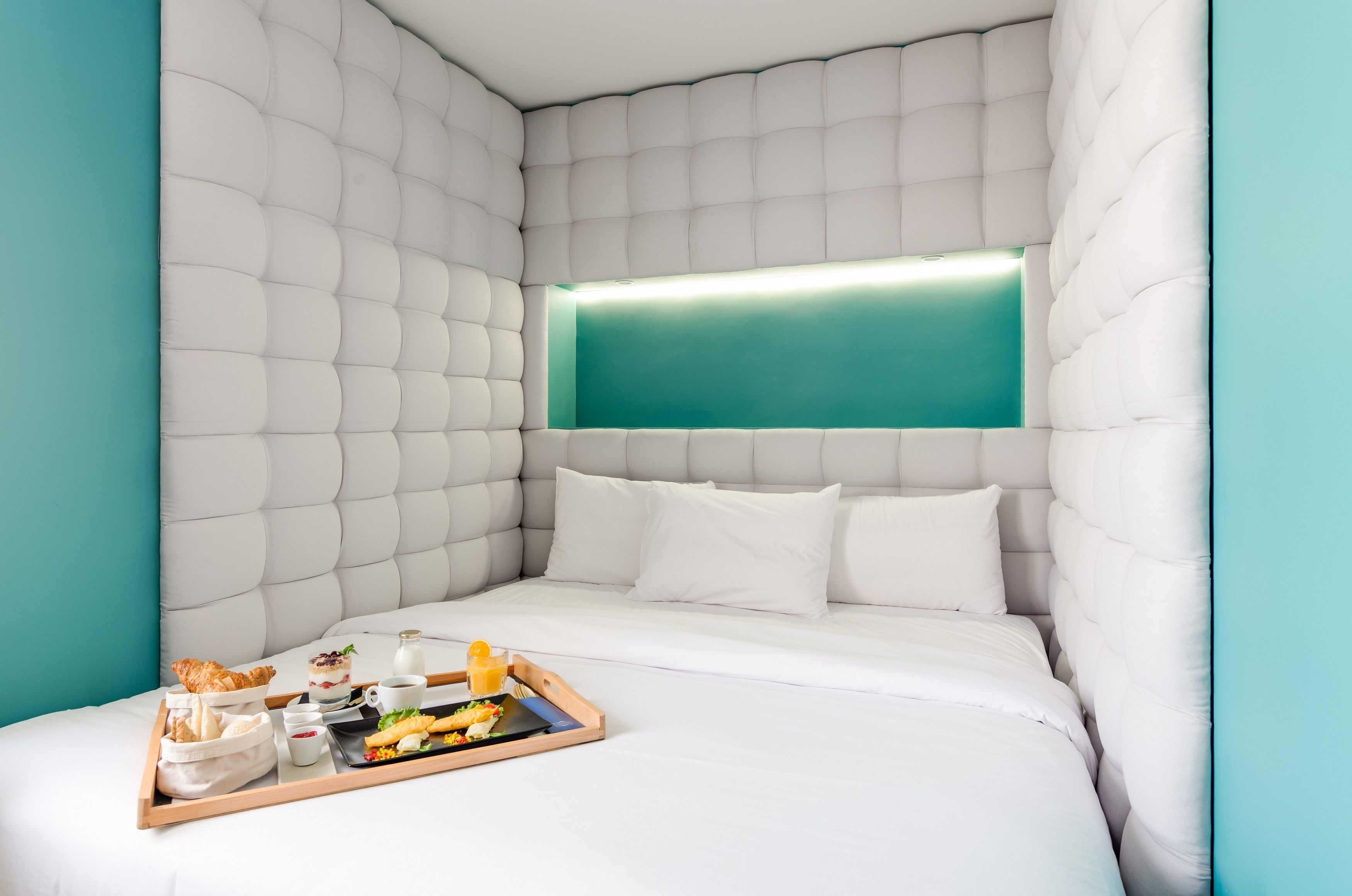 Habitación de hotel moderna con cabecera capitonada, pared de acento color aguamarina y bandeja de desayuno sobre la cama.