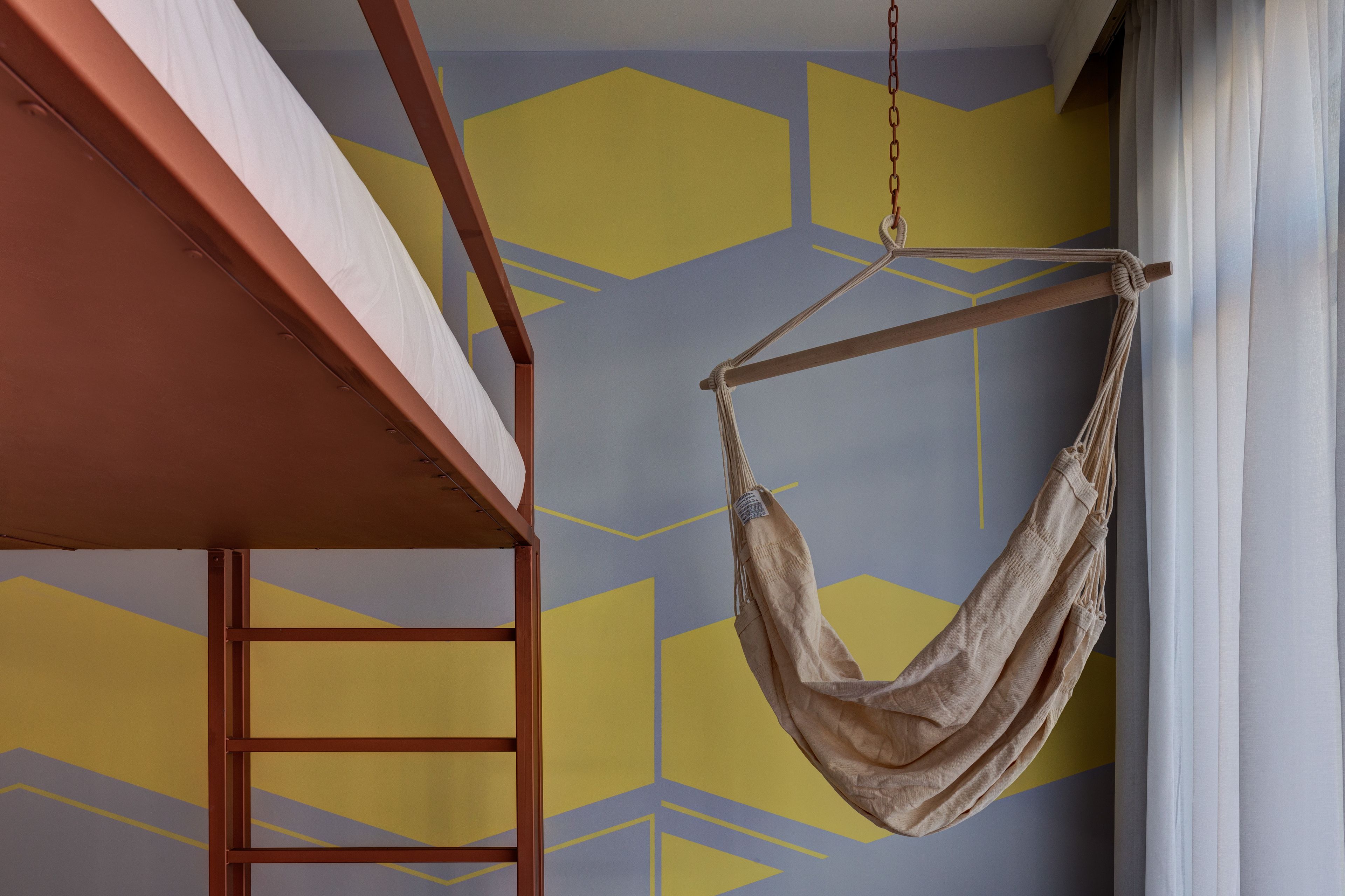 Una hamaca colgando de una viga de madera en una habitación con paredes de color amarillo y gris con diseño geométrico.
