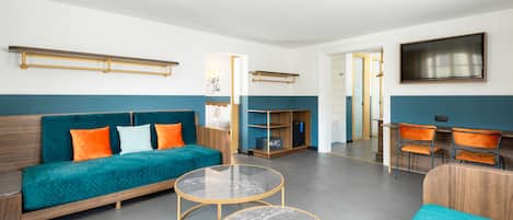 A modern hotel room with a teal sofa, two round tables, a wooden desk, and a TV mounted on the wall.