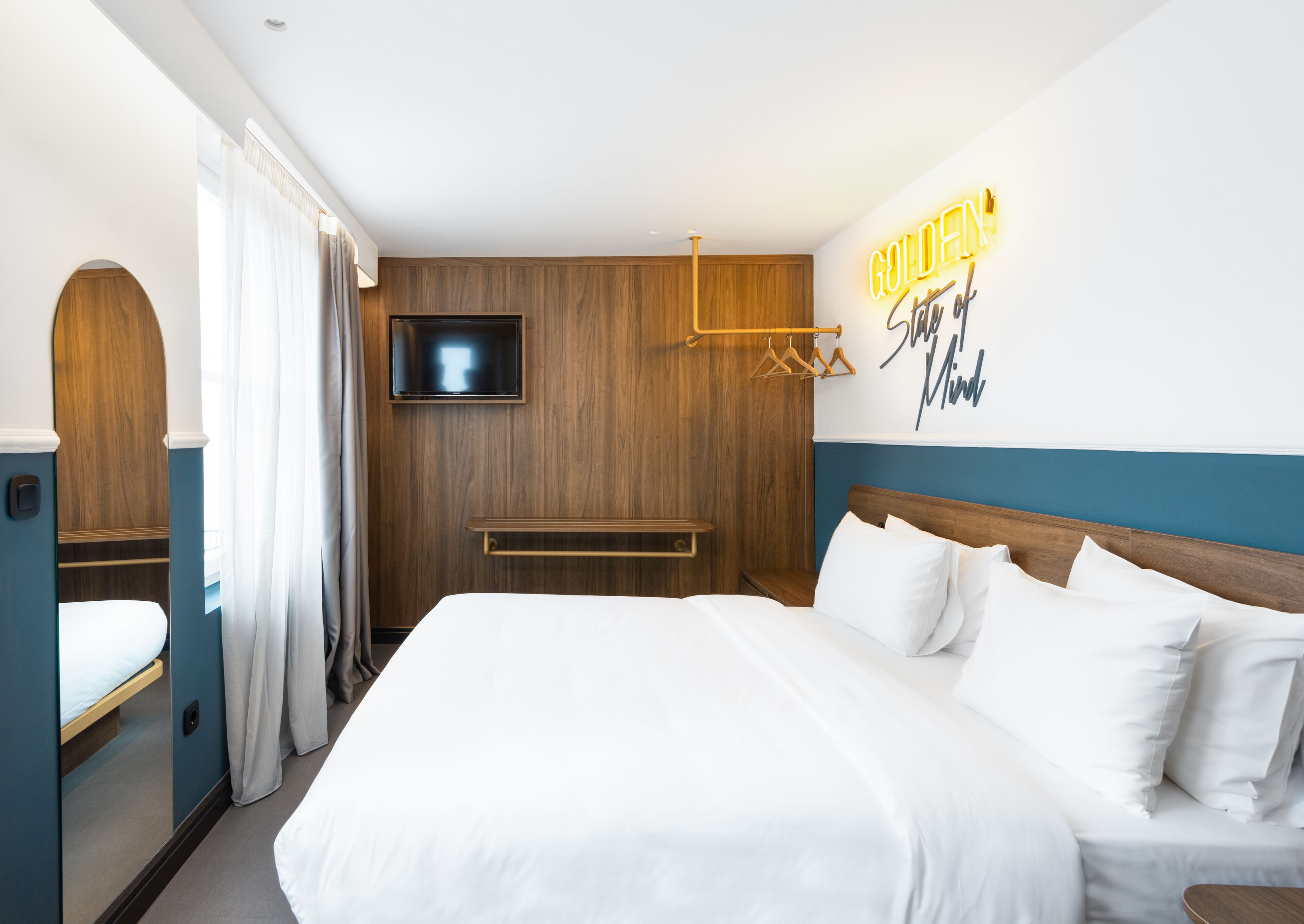 Een hotelkamer met een groot bed, een aan de muur bevestigde televisie, houten lambrisering en een neonbord met de tekst 'GOLDEN State of Mind'.