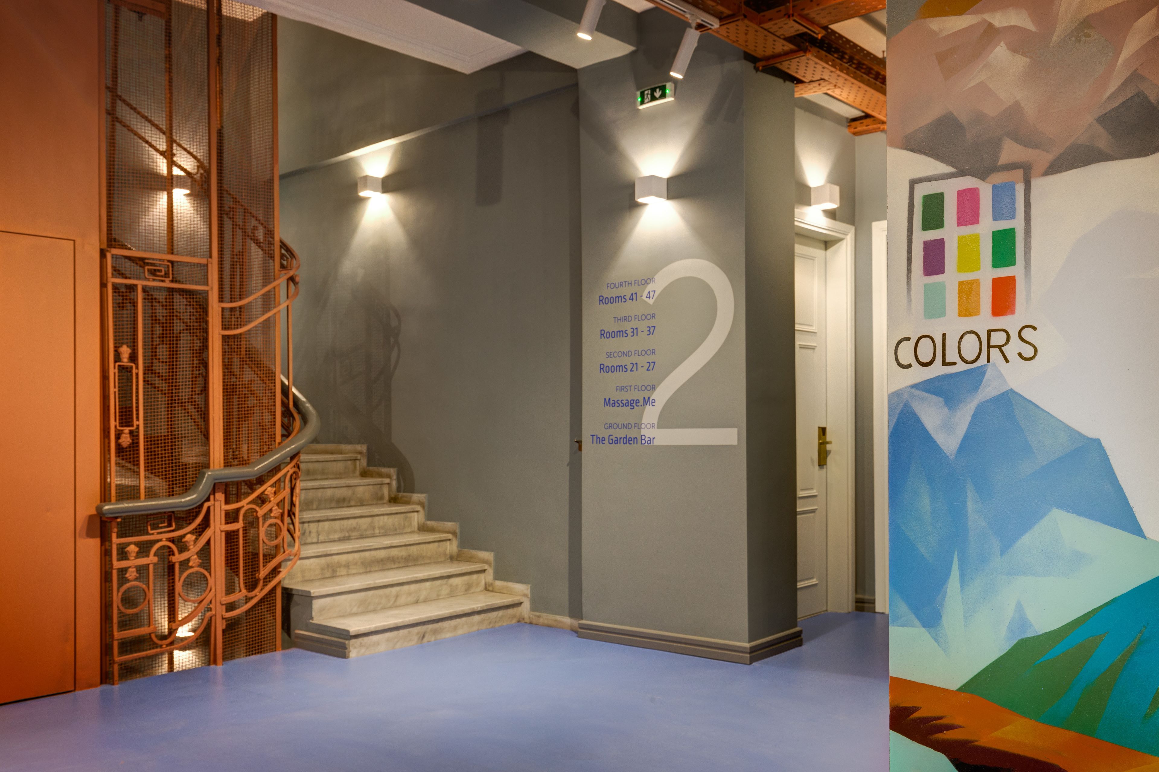 Vestíbulo de hotel con una escalera, una pared con números de habitación y un mural colorido.