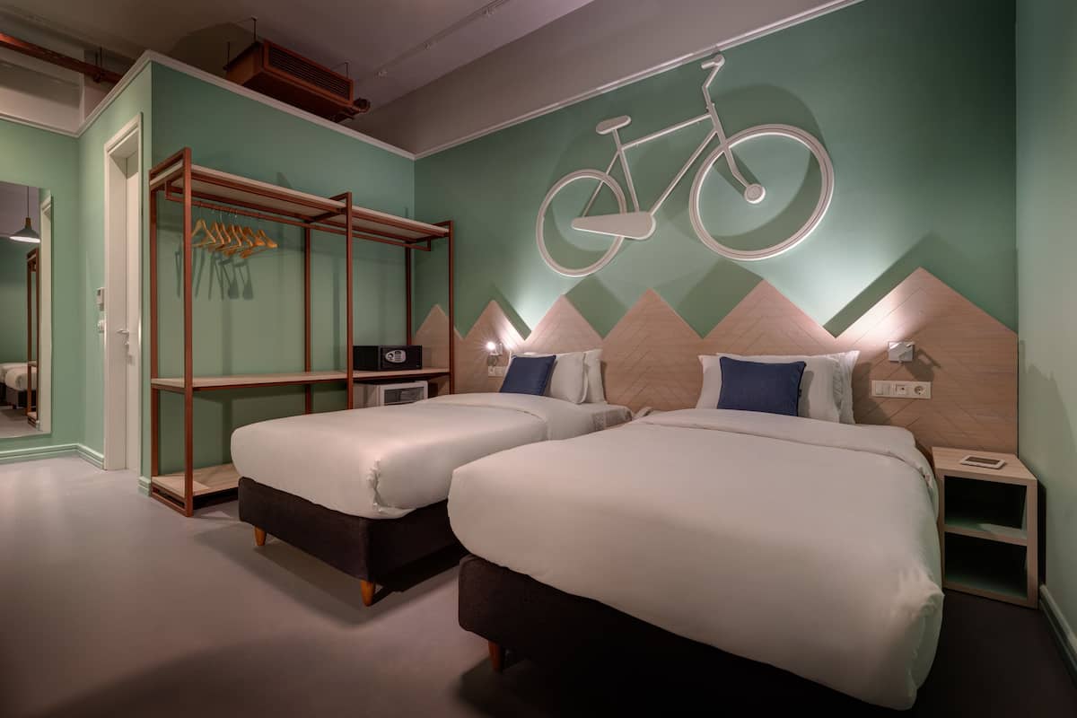 A hotel room with two beds, a wooden wardrobe, a mirror, and a wall-mounted bicycle decoration.