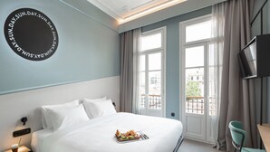 Una cama bien hecha con una bandeja de comida, un televisor montado en la pared y vistas a la ciudad a través de amplias ventanas.