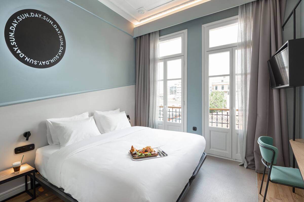 A neatly made bed with a tray of food, a TV mounted on the wall, and a view of the city through large windows.