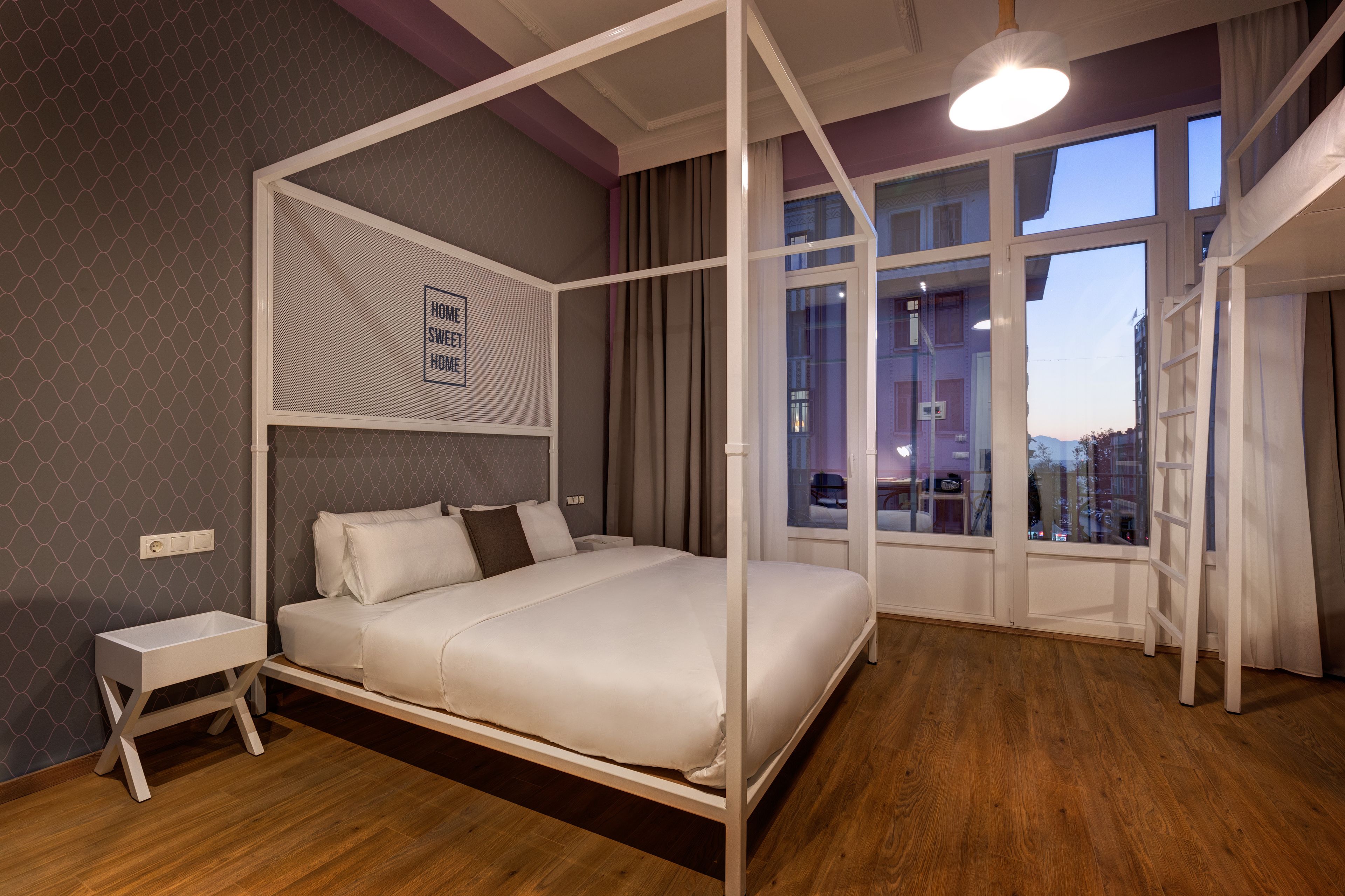 Een moderne slaapkamer met een hemelbed, een klein nachtkastje en een stadszicht door grote ramen.