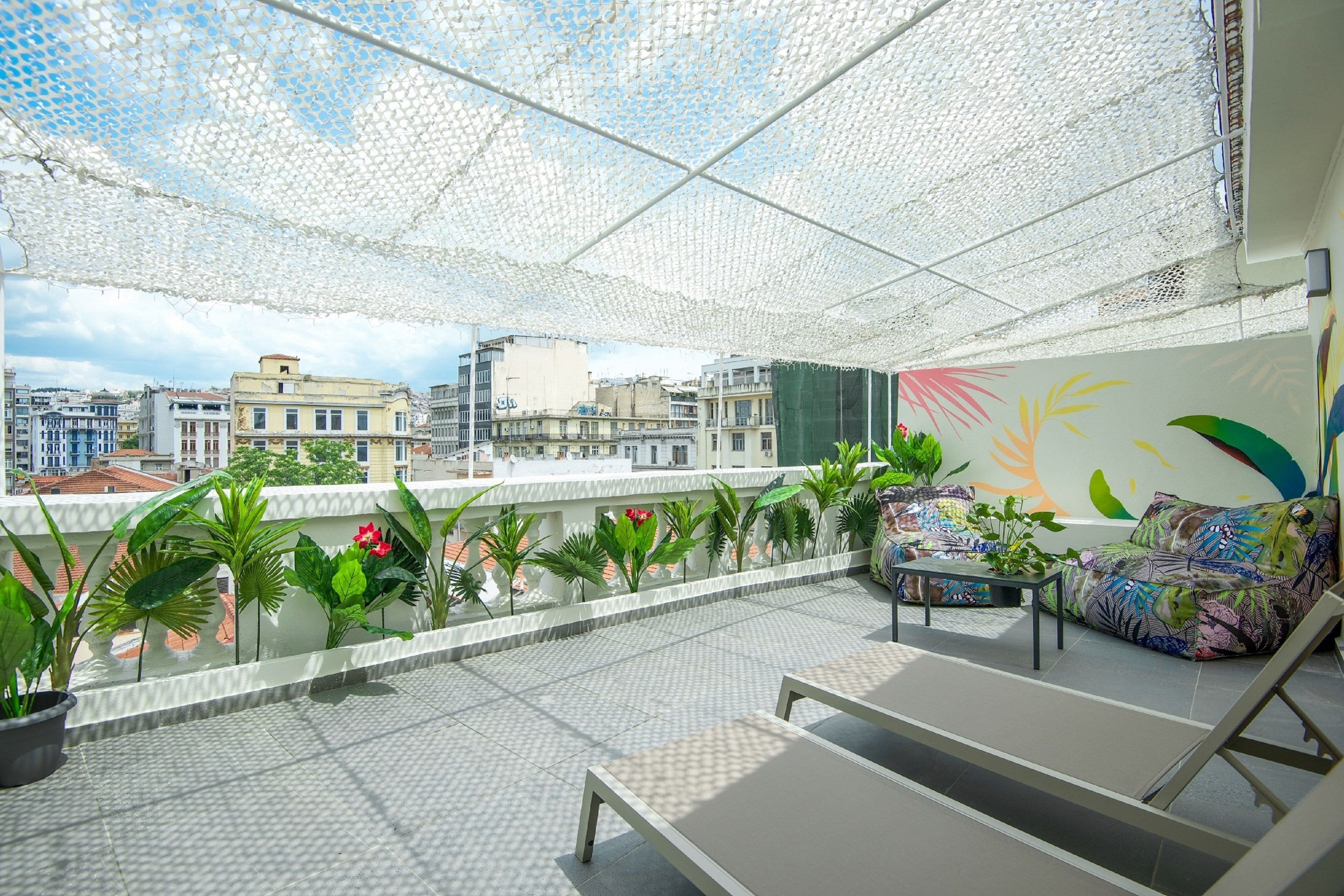 A rooftop terrace with a white mesh canopy, potted plants, a sofa, and a view of city buildings.