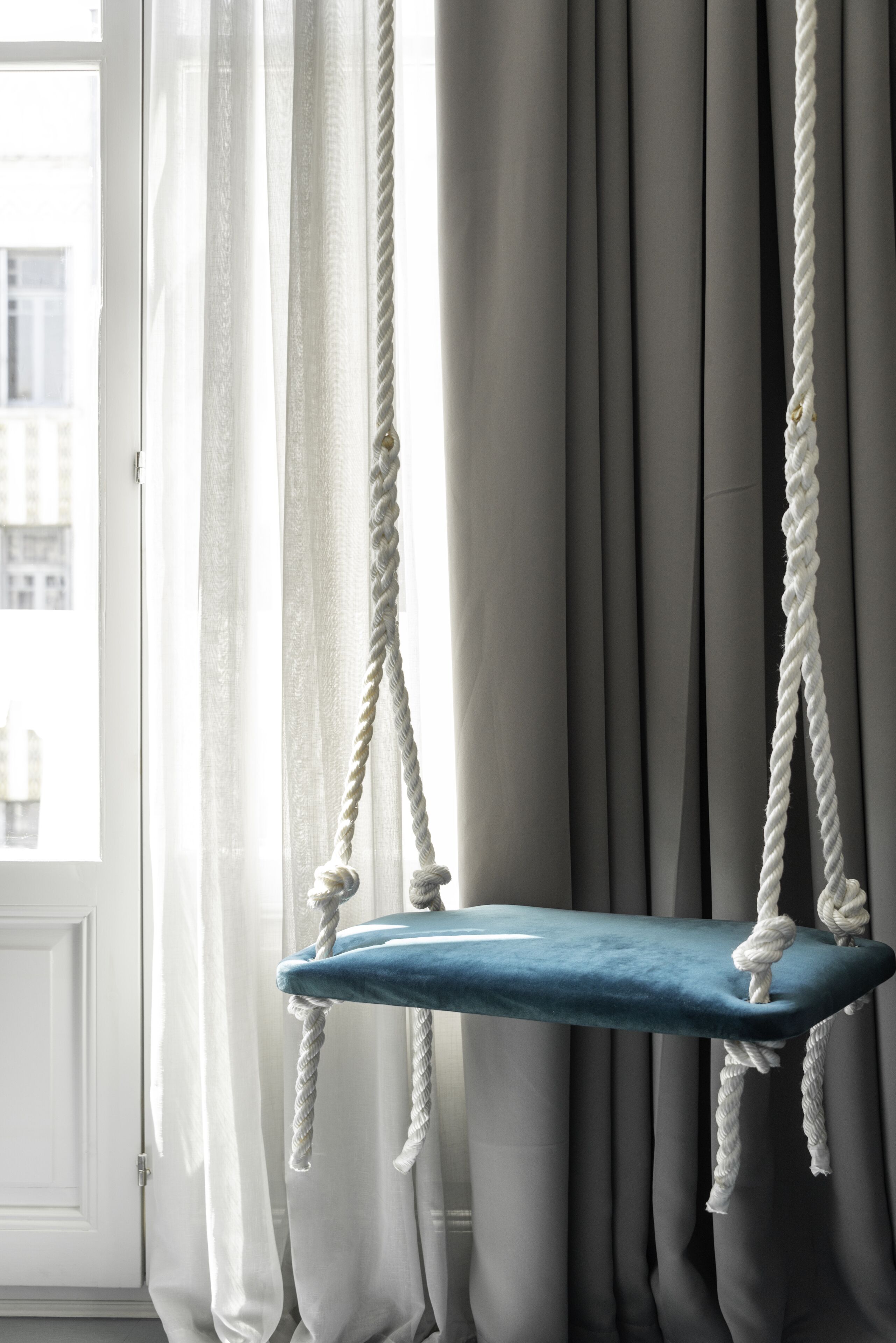 A room with a blue cushioned swing hanging from a rope, grey curtains, and a view of buildings outside.