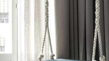 Una habitación con un columpio azul acolchado colgado con una cuerda, cortinas grises y vistas de edificios.