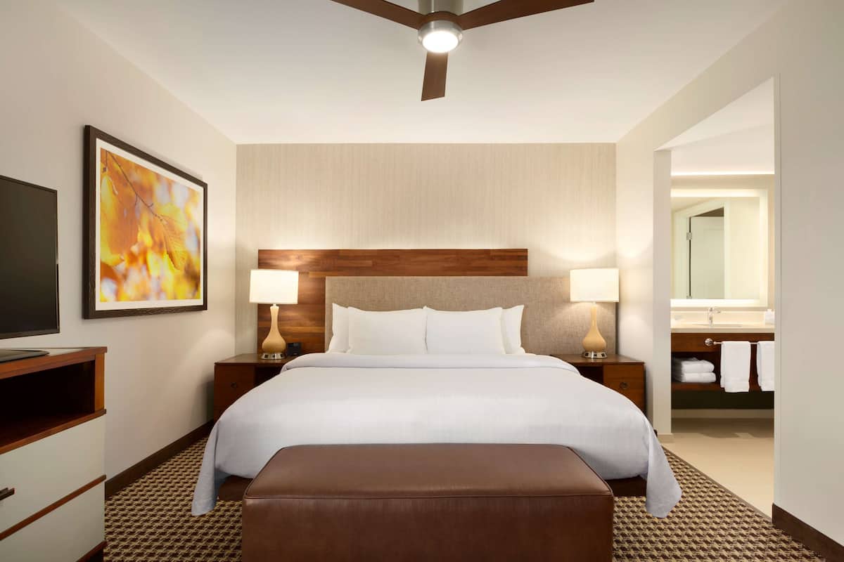 A hotel room with a bed, two bedside lamps, a television, a wooden headboard, a large mirror, and a bathroom with a sink and towel rack.