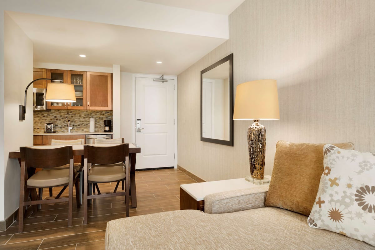 A hotel room with a bed, a small table, two chairs, a kitchen area with cabinets and a microwave, and a large mirror.